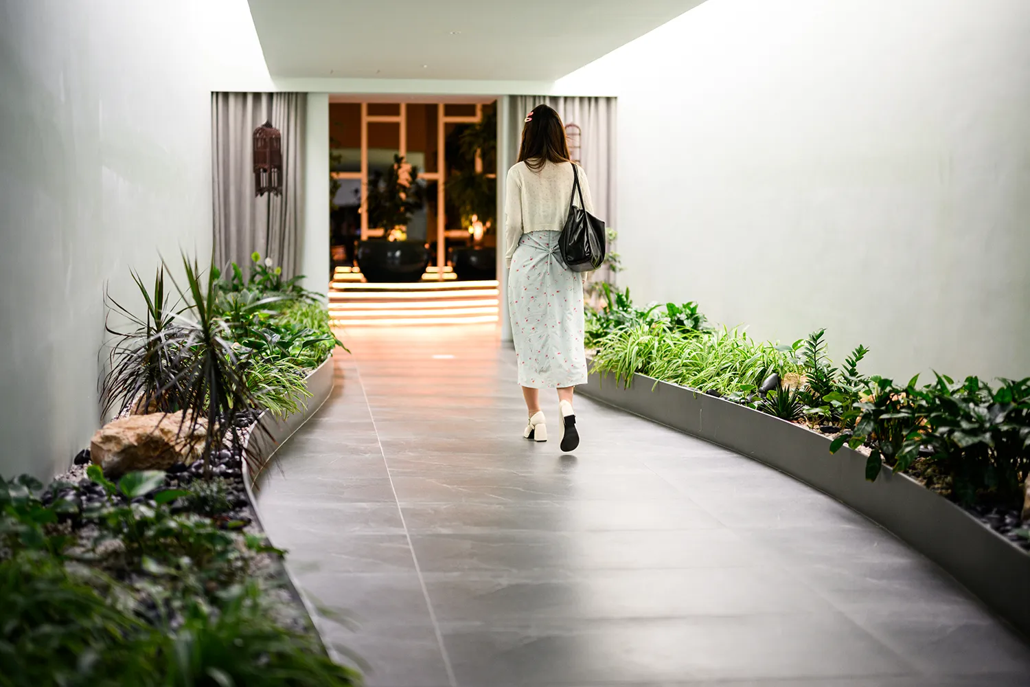 A woman walks down a minimalist, modern indoor hallway lined with lush green plant beds on both sides, leading toward a warm, brightly lit entrance.