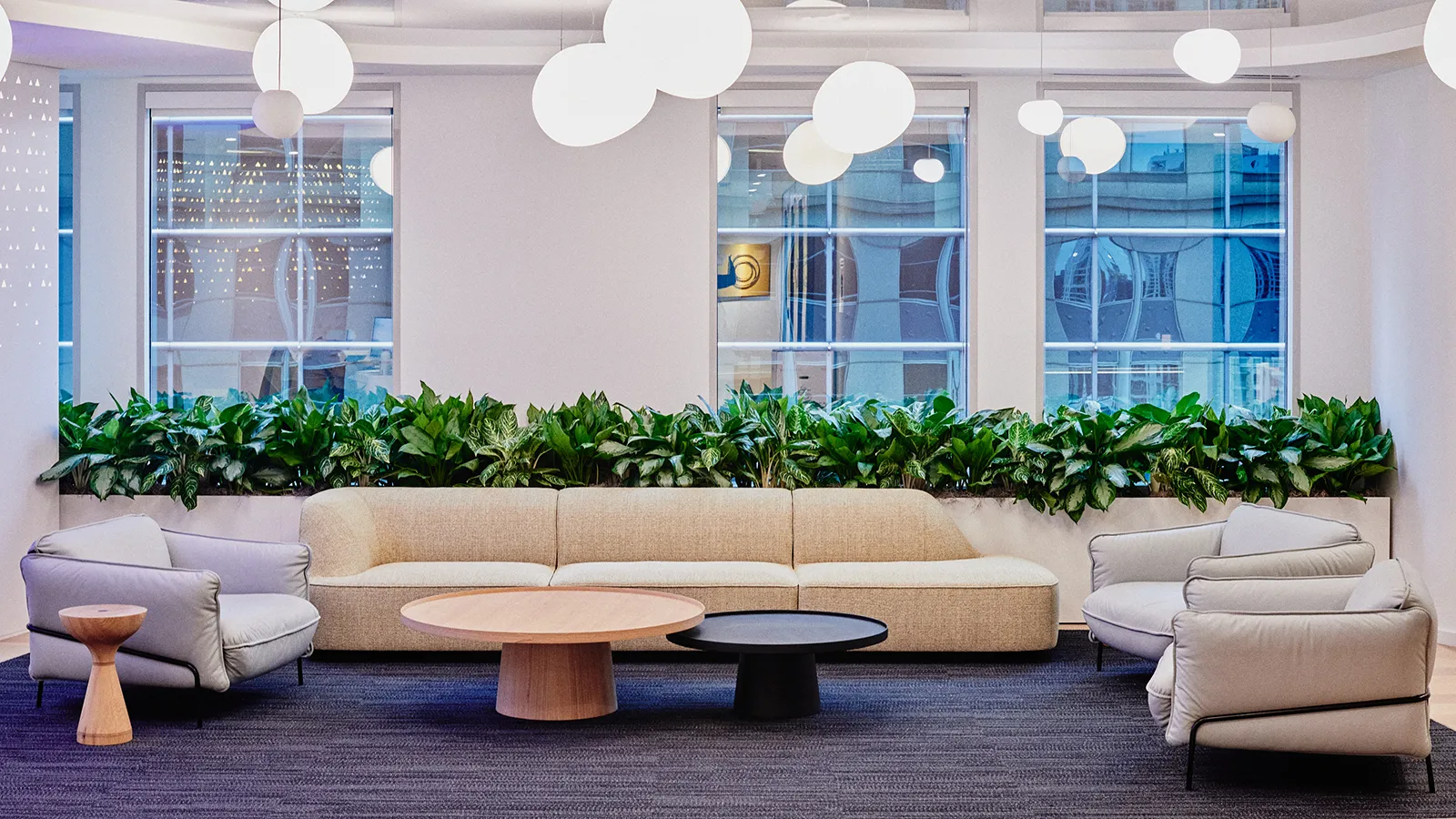 A modern corporate office lounge with extensive lush green indoor plants in a long built-in planter under windows, designed using ecotherapy principles to combat nature deficit disorder for employees.
