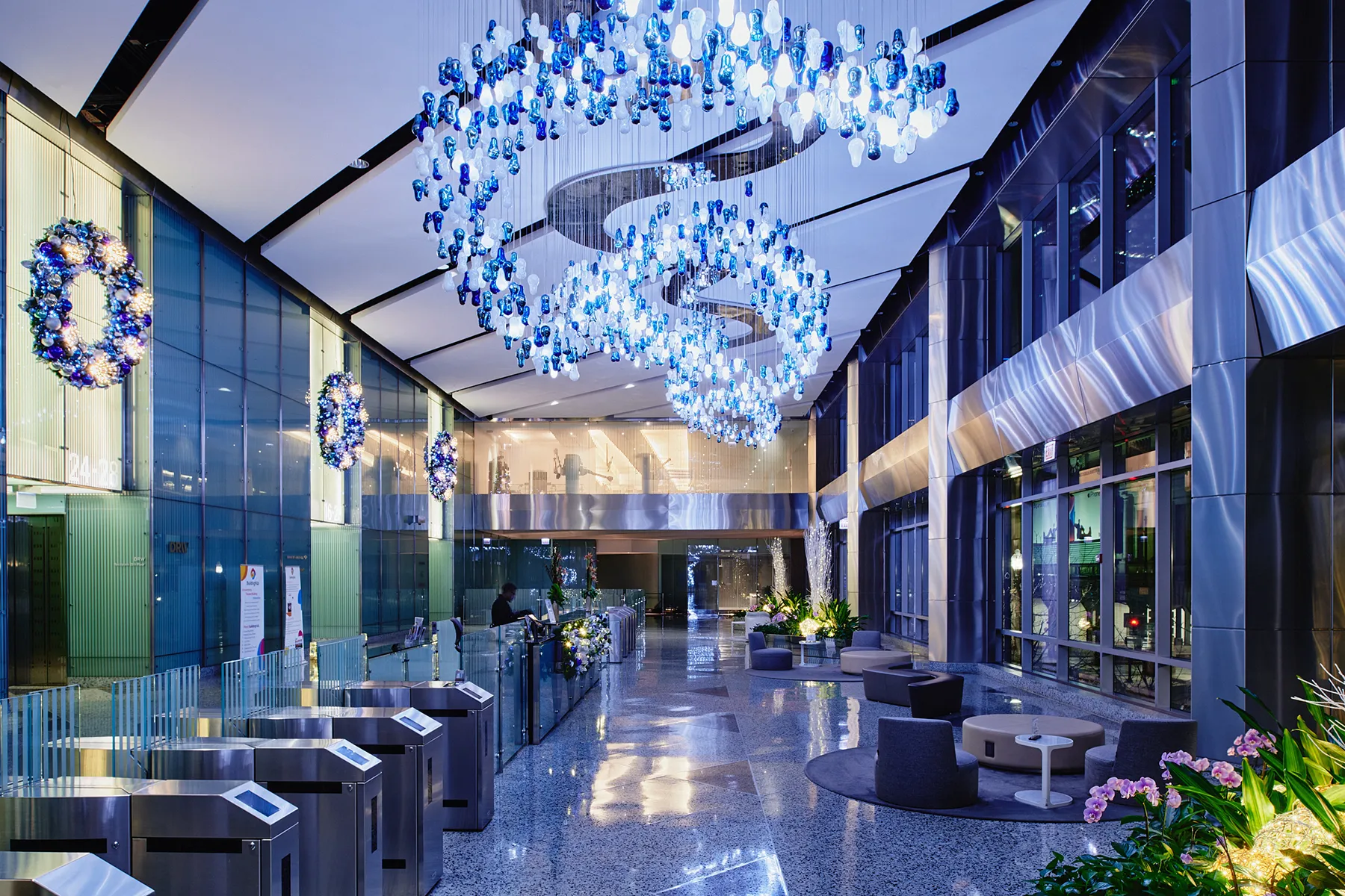 A wide view of a modern corporate lobby featuring grand-scale commercial holiday decor by Amlings, including large suspended wreaths with blue and silver ornaments mounted on an expansive ceiling.