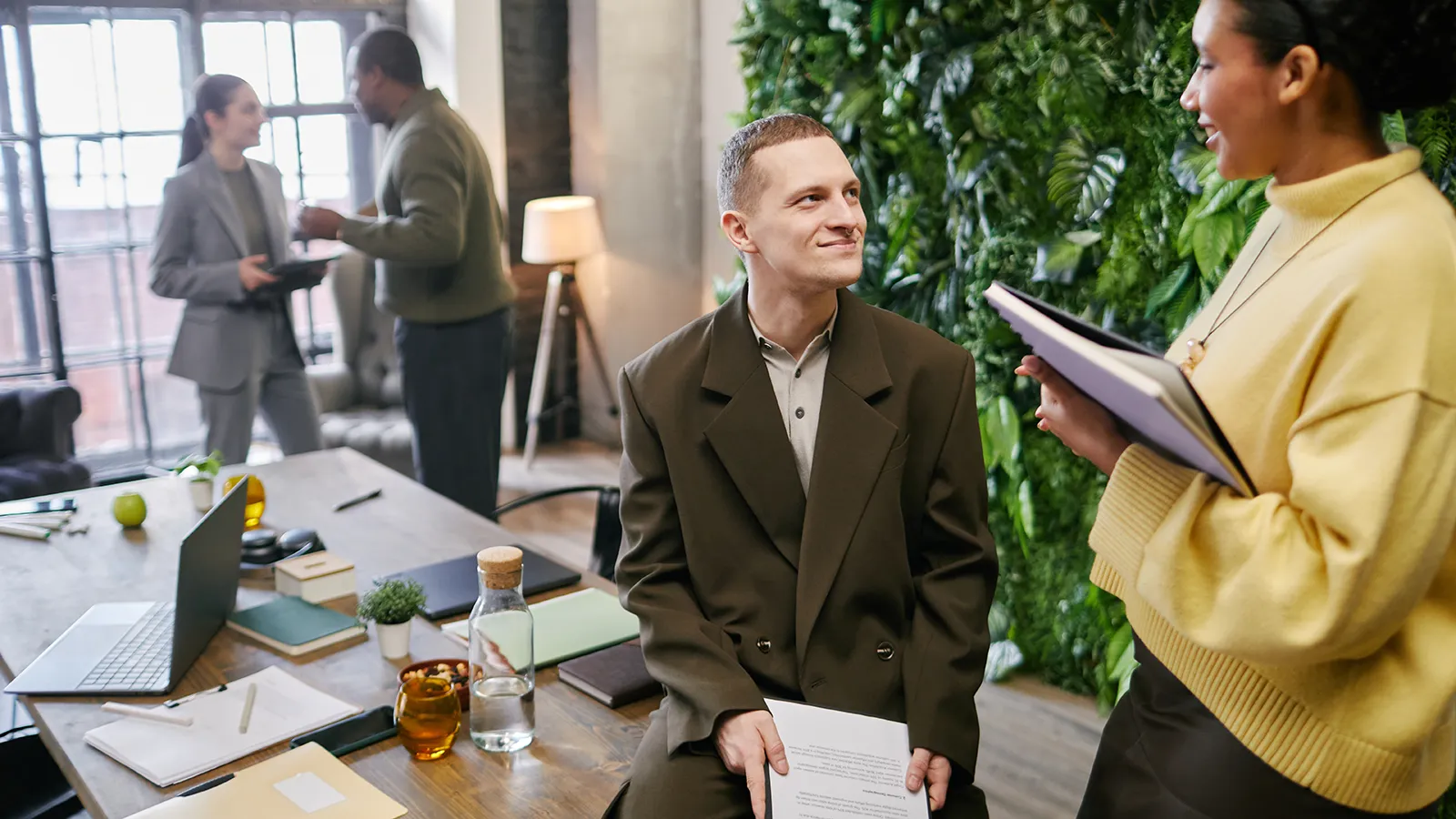 Office employees smile at each other. Behind them is a large, vibrant living wall made of lush green foliage, enhancing the biophilic design of the workplace.