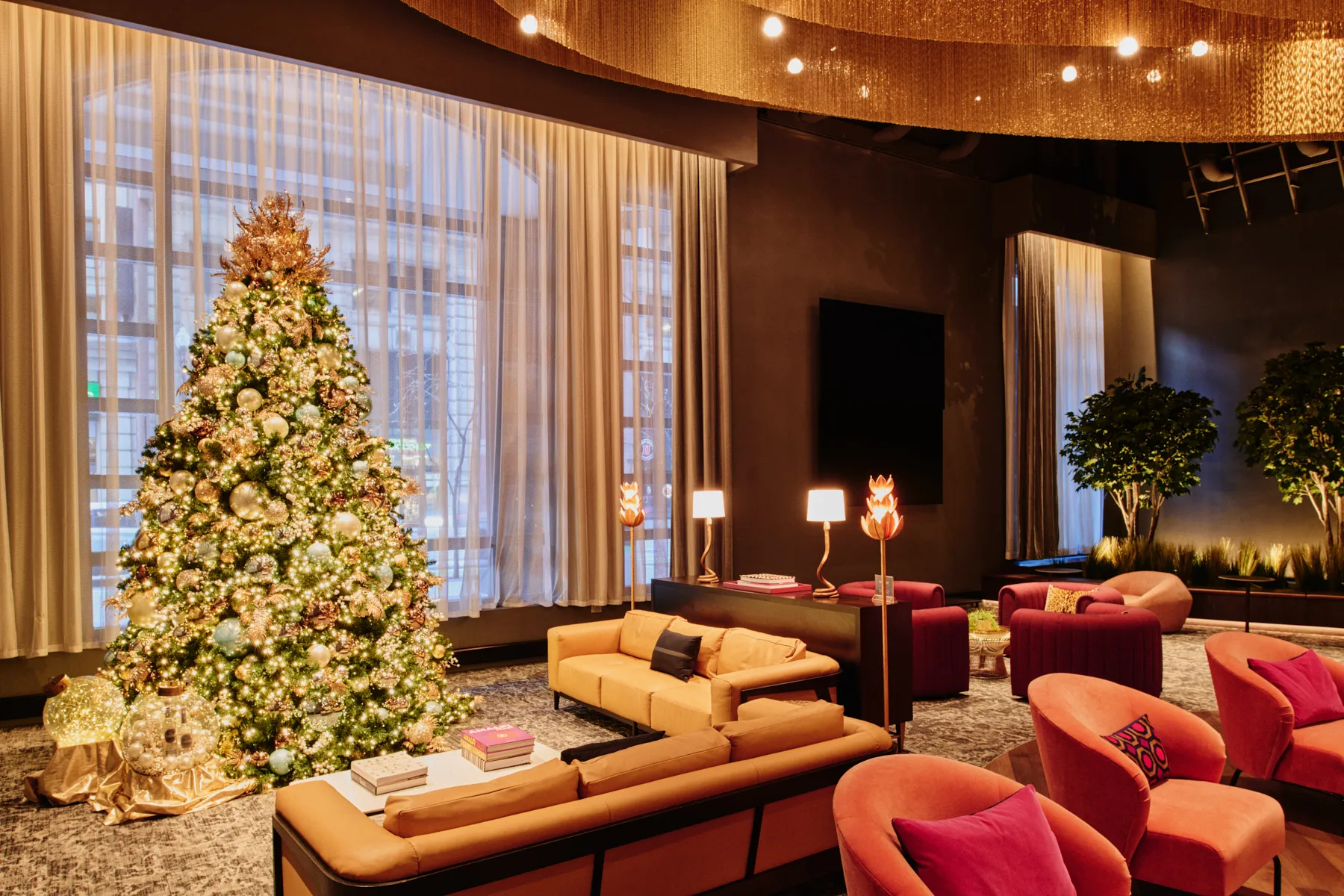 A sophisticated commercial lounge area with a massive, gold-lit Christmas tree, peach and purple velvet armchairs, and warm ceiling fixtures, illustrating luxurious holiday decorations for businesses that create a welcoming 