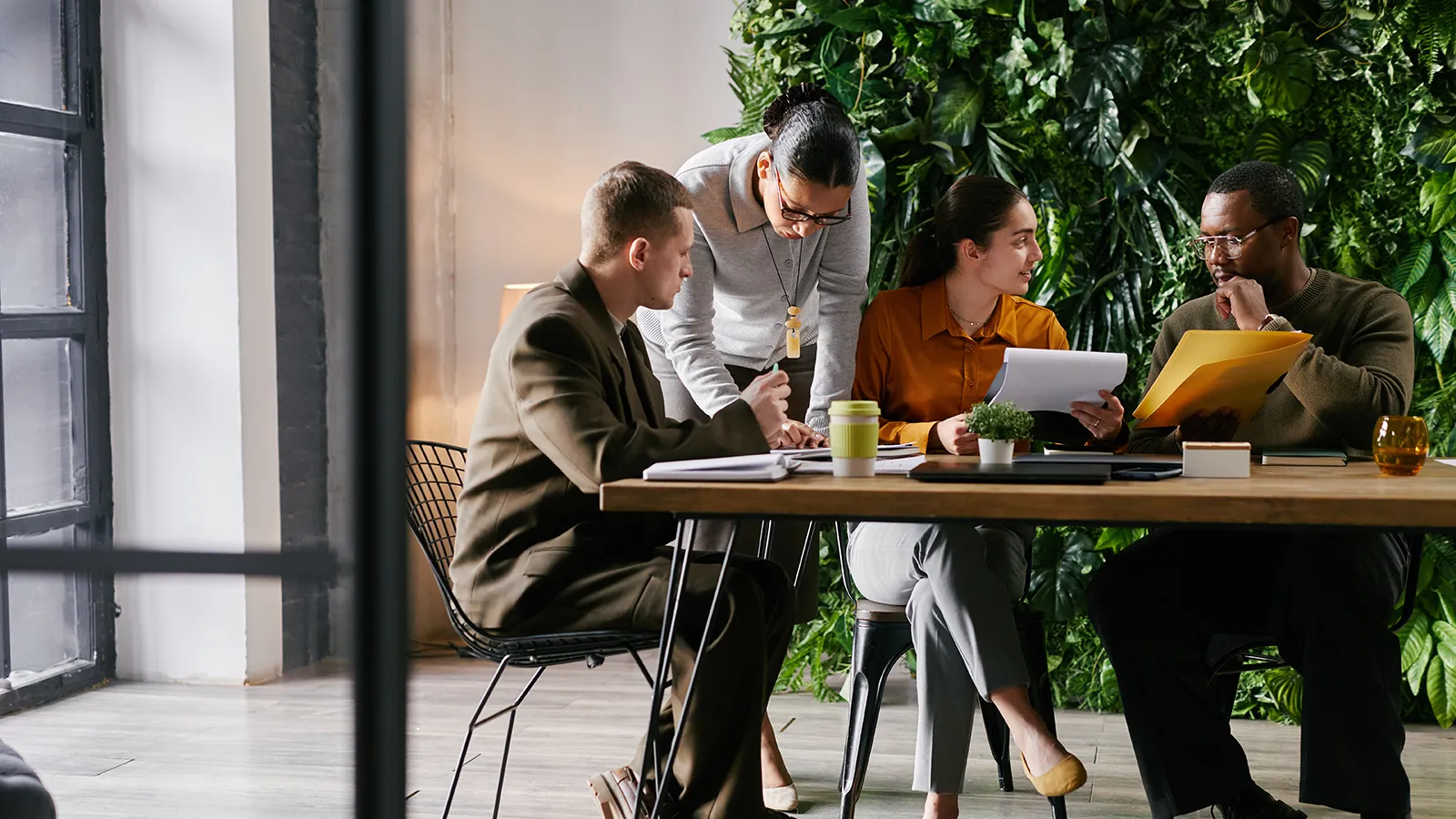 A diverse group of corporate professionals collaborates and takes notes at a meeting table within a modern, naturally lit office environment. A large, thriving vertical garden, a professional living wall installation, covers the entire back wall with diverse green foliage.
