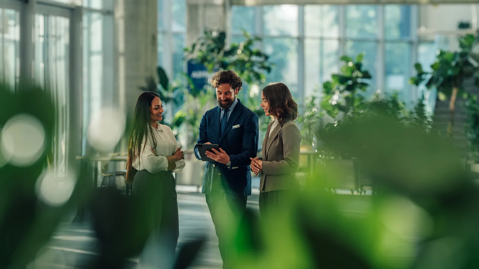 Business professionals using a tablet in a modern office building lobby with indoor greenery and large windows.