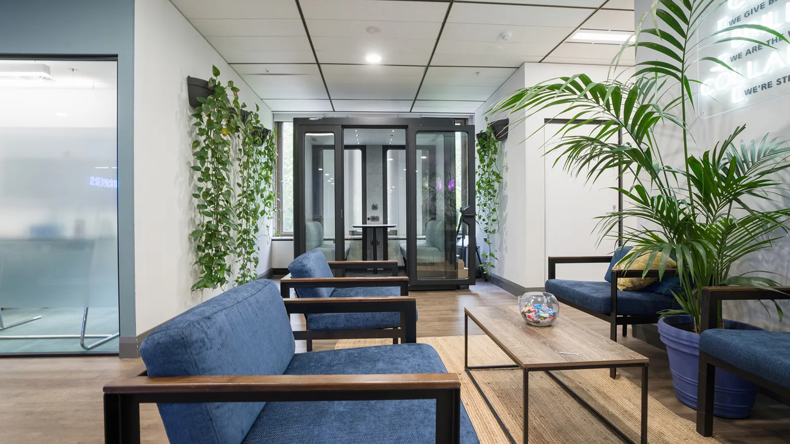 Modern office lounge with blue upholstered seating, green plants, and glass-walled meeting rooms.